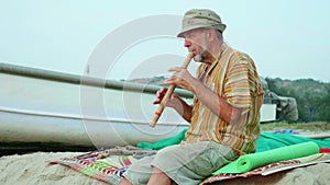 Senior man playing bamboo flute on the beach next to fishing boat