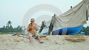 Senior man playing bamboo flute on the beach next to fishing boat