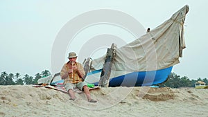 Senior man playing bamboo flute on the beach next to fishing boat