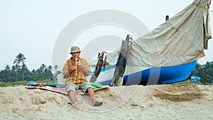 Senior man playing bamboo flute on the beach next to fishing boat