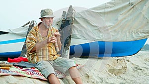 Senior man playing bamboo flute on the beach next to fishing boat