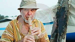 Senior man playing bamboo flute on the beach next to fishing boat