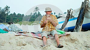 Senior man playing bamboo flute on the beach next to fishing boat