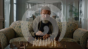 Senior man playing chess alone in classic living room. Focused mature gentleman