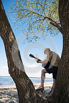 Senior man playing guitar while standing on big tree at riverside