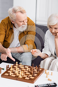 Senior man pointing at figures on chess board near wife and remote controller on blurred foreground.