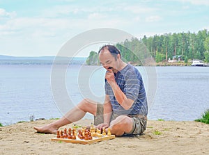 Senior man pondering a move in a game of chess and sitting on the beach near the lake