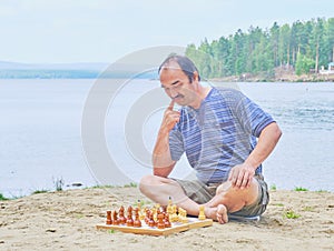 Senior man pondering a move in a game of chess and sitting on the beach near the lake
