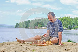 Senior man pondering a move in a game of chess and sitting on the beach near the lake