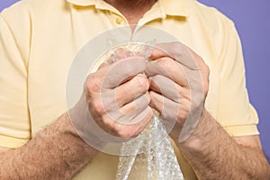 Senior man popping bubble wrap on light purple background, closeup. Stress relief