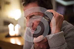 Senior man praying, holding Bible in his hands, eyes closed.