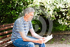 Senior man reading a book on a bench in the park in bright sunny day. Copy space