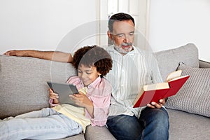 Senior Man Reading Book While Grandson Using Digital Tablet Indoor