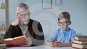 Senior man reading and looking at his grandson playing games on phone, leisure