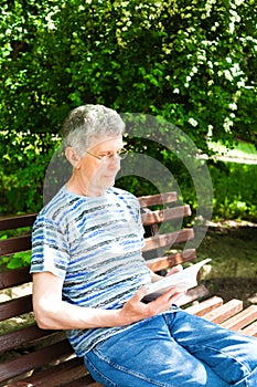 Senior man is resting on a bench in the park and reading a book. A bright sunny day