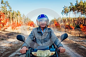 Senior man riding scooter on autumn forest road. Driver in helmet riding moped