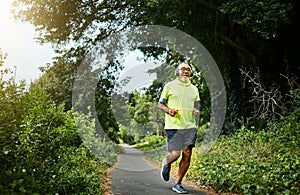 Senior man, running and listening to music on road in nature, countryside or forest for exercise in retirement. Fitness