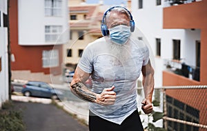 Senior man running outdoor while wearing face protective mask - Mature male doing jogging listening music with headphones during