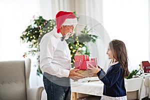 A senior man with a Santa hat giving presents to a small girl at Christmas time.