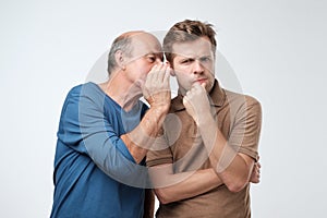 Senior man sharing secret or whispering gossips into his son ear. Telling family secret concept