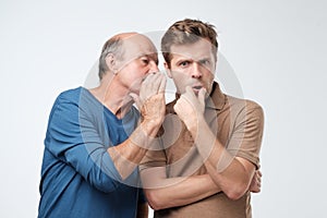 Senior man sharing secret or whispering gossips into his son ear. Telling family secret concept