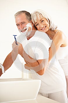 Senior Man Shaving In Bathroom Mirror With Wife