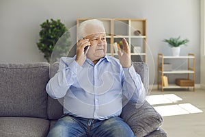 Senior man sits on a sofa in a bright living room and talks on the phone with his family.
