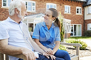 Senior Man Sitting On Bench And Talking With Nurse In Retirement Home