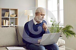 Senior man sitting on couch at home, working on his laptop computer and thinking