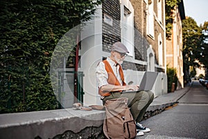 Senior man sitting on curb working on his laptop.