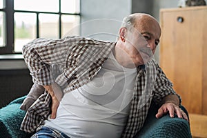 Senior man sitting indoor on the armchair feeling sudden ache in his back