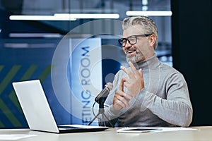 Senior man sitting an office with a microphone in front of a laptop. Talks on a video call