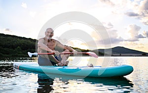 Senior man sitting on paddleboard on lake in summer. Copy space.