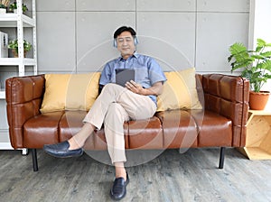 A senior man sitting on sofa and listening to music via headphones with wireless tablet computer in hand with happiness manner.