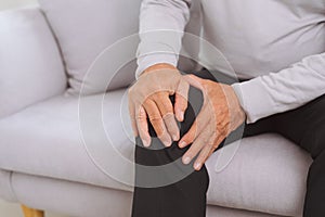 Senior man sitting on a sofa in the living room at home and touching his knee by the pain