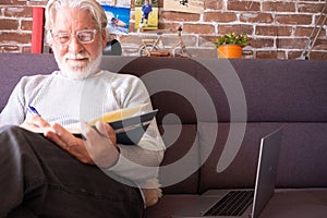 Senior man sitting on sofa and reading book at home - retired, leisure and people concept