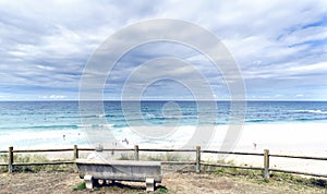 Senior man sitting on a stone bench looking at the atlantic ocean on Razo beach and some people on the beach with a sky with many