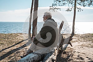 Senior man sitting at the tree log at the seashore and looking at the water surface