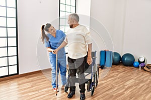 Senior man sitting on wheelchair being assited by geriatric nurse, handicapped mature man getting help to stand up