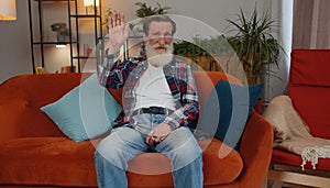 Senior man smiling friendly at camera, waving hands gesturing hello, hi, greeting at home on couch