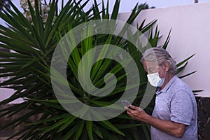 Senior man standing against  a wall and a big green plant with face mask because of coronavirus - using smart phone - concept of