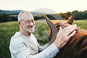 A senior man standing close to a horse outdoors in nature, stroking it.