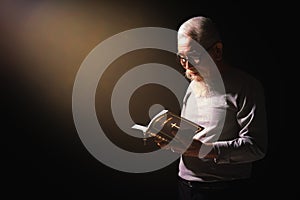 Senior man standing in dark background holding and reading holy bible