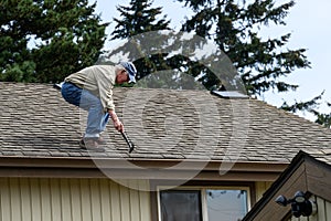 Senior man standing on a house roof with a hammer, pounding down nails that have popped out