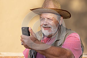 Senior man in straw hat listening music from cellular over headphones while sitting outdoor against clay wall
