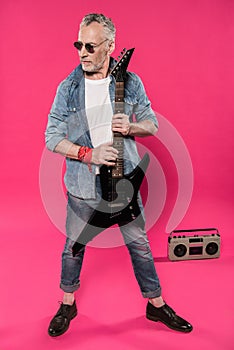 Senior man in sunglasses and denim jacket holding electric guitar and looking away