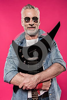 Senior man in sunglasses and denim jacket holding electric guitar and smiling at camera