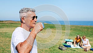 Senior man with sunglasses drinking coffee outdoors with his family in background