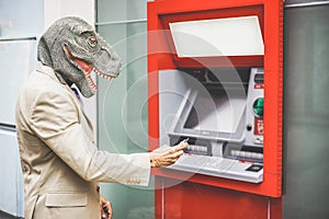 Senior man with t-rex mask withdraw money from bank cash machine with debit card - Crazy new trends advertising concept - Focus on