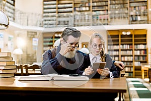 Senior man teacher with his little student pupil girl is using a digital tablet, sitting together at the table in old